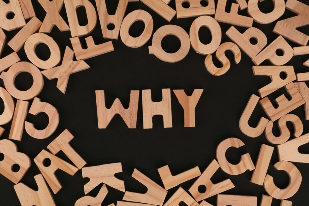 Wooden letters scattered on a black background frame the word “WHY” in the center, spelled in bold wooden block letters, emphasizing the central question of purpose.