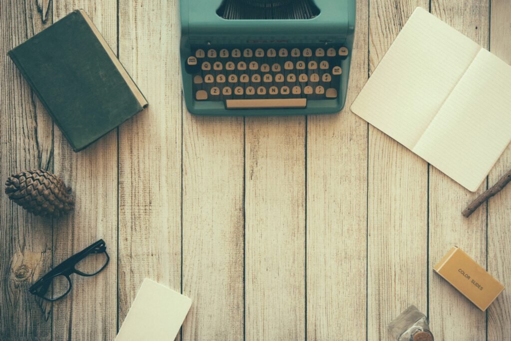 Flat lay of a vintage teal typewriter, open notebook, glasses, and writing accessories on a wooden surface.