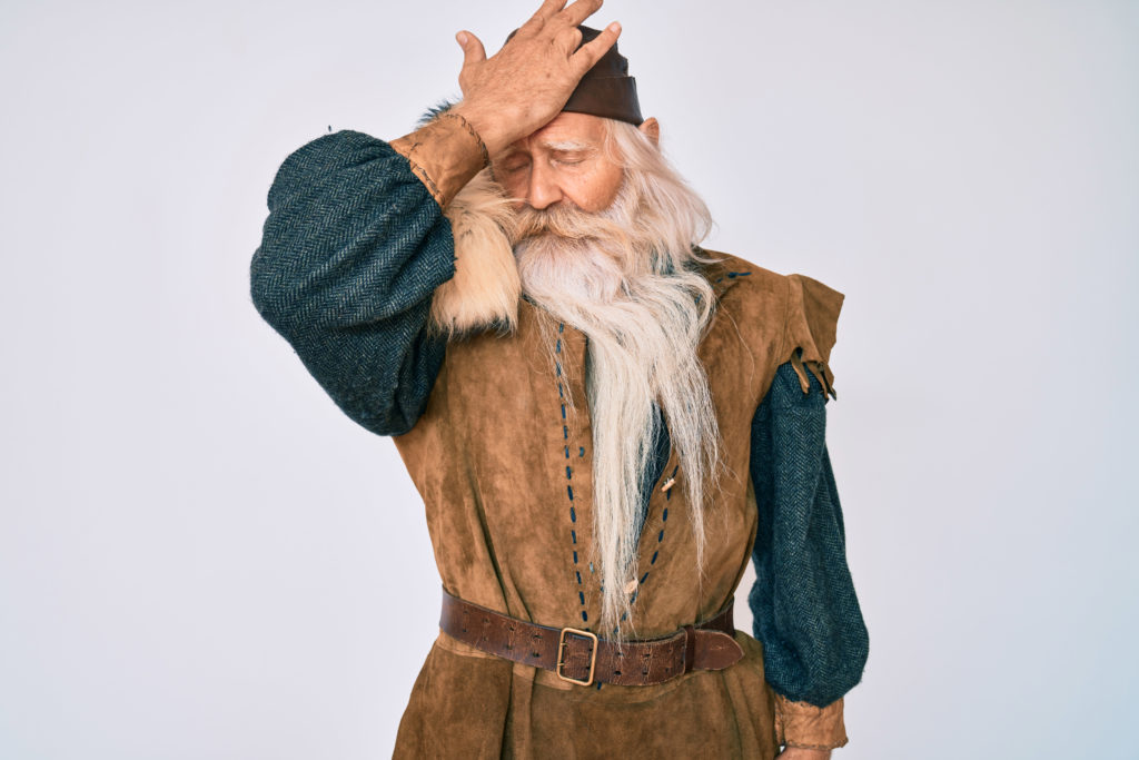Old senior man with grey hair and long beard wearing viking traditional costume surprised with hand on head for mistake, remember error. forgot, bad memory concept.